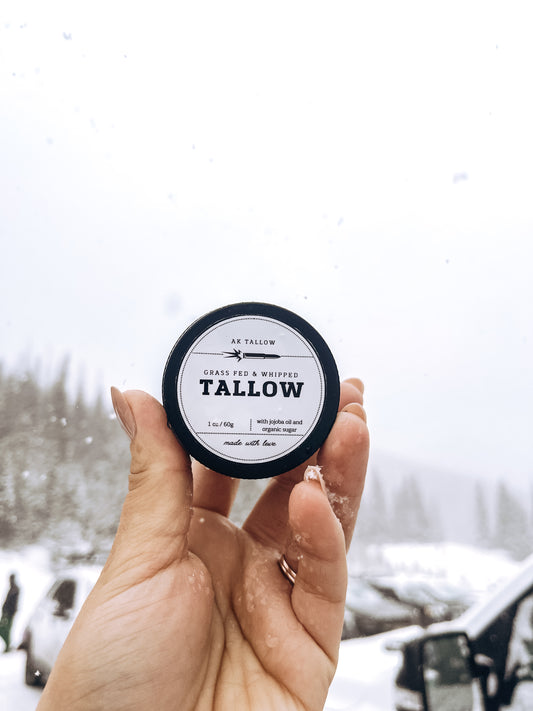 An image of person's hand holding a jar of grass-fed whipped tallow with jojoba oil against a snowy background in the Rocky Mountains