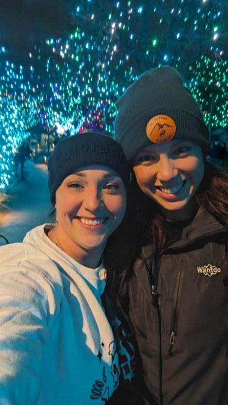 Two smiling women wearing beanies at Denver Zoo light night event with blue christmas lights in backround